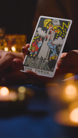 Vertical-Video-Close-Up-Of-Woman-Giving-Tarot-Card-Reading-On-Candlelit-Table-Holding-The-Tower-Card-2